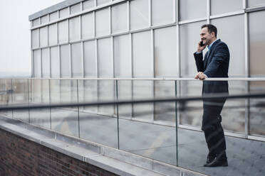 Smiling businessman talking on smart phone by glass railing on balcony - MOEF04021