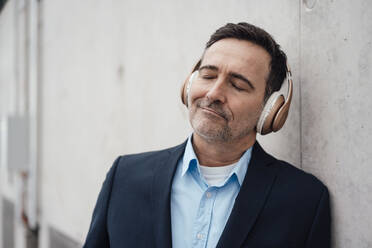 Smiling businessman with eyes closed listening music through wireless headphones by wall - MOEF04037