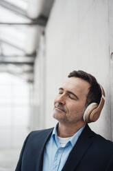 Businessman with eyes closed listening music through wireless headphones by wall - MOEF04038