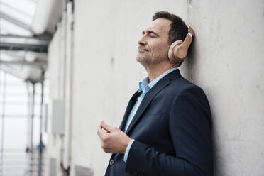Smiling businessman enjoying music through wireless headphones by wall in greenhouse - MOEF04039