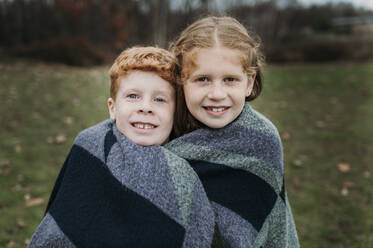 Smiling girl and brother wrapped in blanket on meadow - ELEF00039