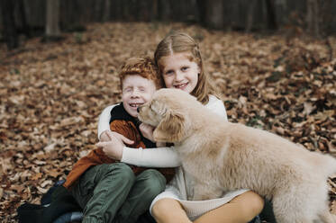 Puppy licking boy's face sitting with sister on autumn leaves in forest - ELEF00042