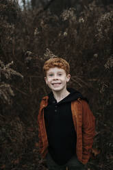Smiling boy standing with hands in pockets in forest - ELEF00048