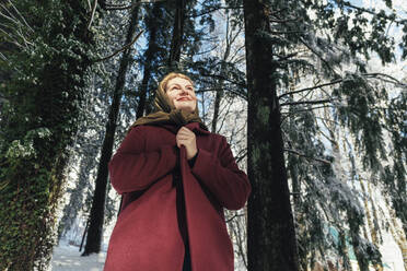 Lächelnde reife Frau vor Bäumen im Winterwald stehend - OMIF00643