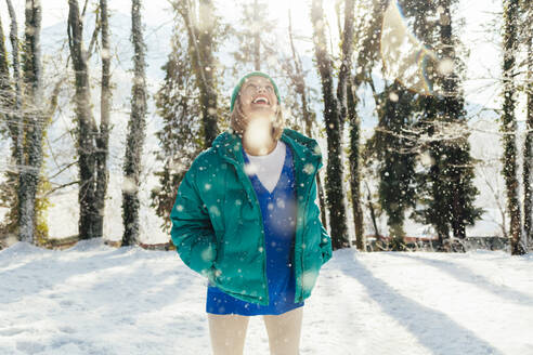 Cheerful woman with hands in pockets enjoying snowfall at forest - OMIF00650