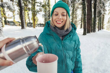 Woman pouring water in mug being held by friend at winter forest - OMIF00652