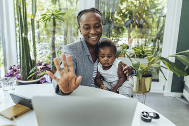 Smiling freelancer with baby boy waving on video call at home - MFF08416