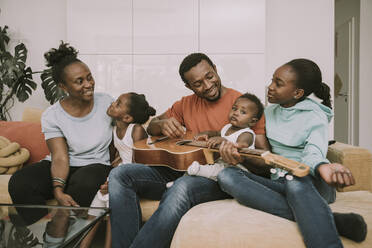 Smiling father playing guitar by family in living room - MFF08444