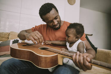 Father showing guitar to baby boy in living room - MFF08448