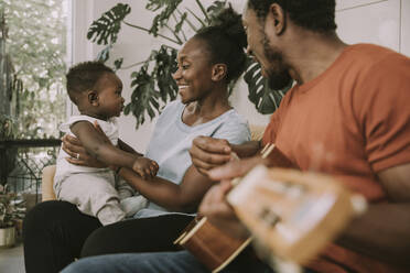 Happy mother with son sitting by father playing guitar at home - MFF08449