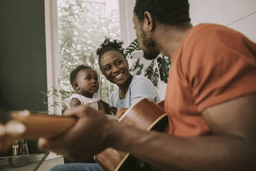 Father plucking guitar by son and mother in living room - MFF08450