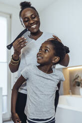 Lächelnde Mutter kämmt die Haare ihrer Tochter im Badezimmer zu Hause - MFF08471