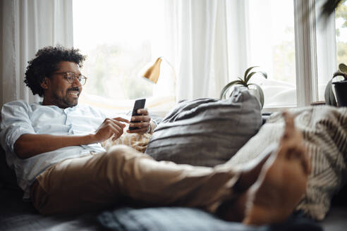 Glücklicher Mann, der ein Smartphone benutzt und zu Hause auf dem Sofa sitzt - GUSF07106