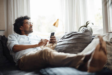 Nachdenklicher Mann mit Smartphone sitzt zu Hause auf dem Sofa - GUSF07107