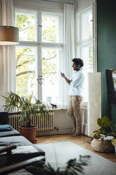 Happy man using mobile phone and listening music through wireless headphones by window at home - GUSF07123