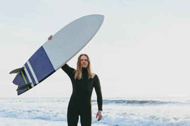 Mann mit erhobener Hand hält Surfbrett am Strand - OMIF00663