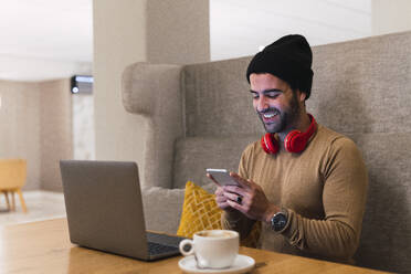 Glücklicher Geschäftsmann mit Strickmütze, der in einem Café sein Smartphone benutzt - PNAF03265