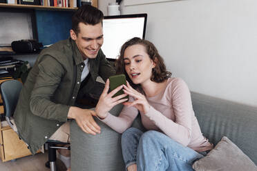 Junge Frau teilt ihr Smartphone mit ihrem Freund, der auf einem Stuhl im Wohnzimmer sitzt - VPIF05345