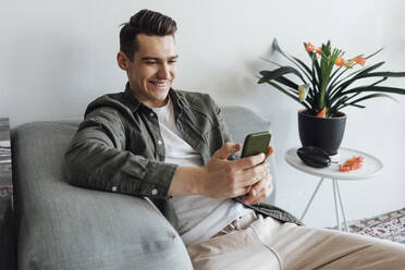 Glücklicher junger Mann, der zu Hause mit seinem Mobiltelefon im Internet surft - VPIF05352