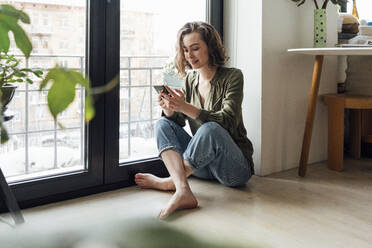 Junge Frau, die mit ihrem Mobiltelefon durch ein Glasfenster zu Hause im Internet surft - VPIF05364