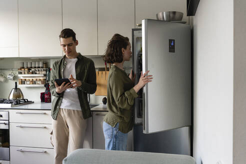 Young man using tablet PC by woman opening refrigerator in kitchen at home - VPIF05370