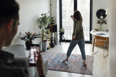 Young man photographing girlfriend singing through smart phone in living room - VPIF05373