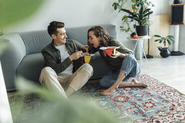 Happy young man looking at girlfriend eating noodles in living room - VPIF05380