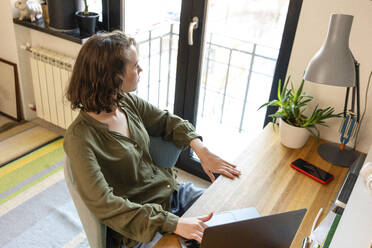 Young woman with laptop looking through window sitting at home - VPIF05399