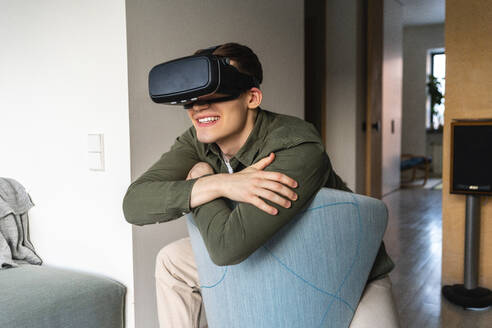 Glücklicher junger Mann, der einen Virtual-Reality-Simulator trägt und sich zu Hause an einen Stuhl lehnt - VPIF05425