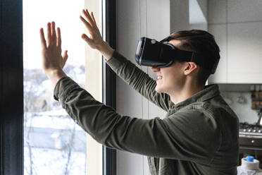 Smiling young man wearing virtual reality simulator touching glass window at home - VPIF05427