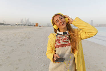 Glückliche Frau in gelber Jacke hört Musik über drahtlose Kopfhörer am Strand - TYF00083