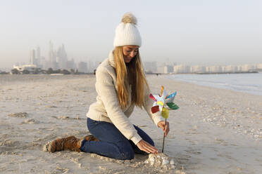 Happy woman sitting on sand with multi colored pinwheel toy at beach - TYF00089