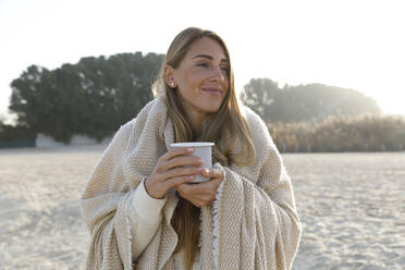 Lächelnde Frau in Decke hält Teetasse am Strand - TYF00093