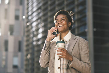 Cheerful businesswoman holding coffee cup talking through mobile phone on sunny day - AGOF00240