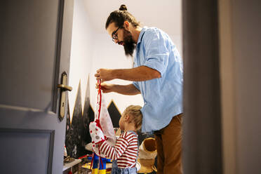 Father helping son putting on apron at home - ASGF02138