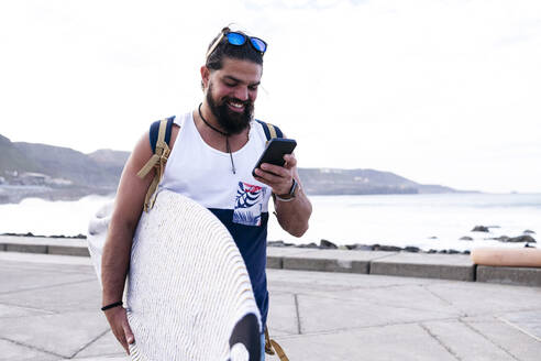 Happy man with surfboard using smart phone by sea - ASGF02149