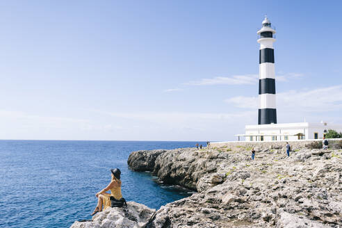 Frau sitzt auf einem Felsen und betrachtet das Meer an einem sonnigen Tag, Leuchtturm von Artrutx, Menorca, Spanien - DGOF02284