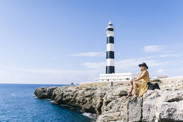 Frau sitzt auf einem Felsen am Meer beim Leuchtturm von Artrutx auf Menorca, Spanien - DGOF02285