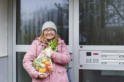 Smiling senior woman with vegetables and fruits in mesh bag at entrance of apartment - IHF00762