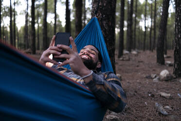 Happy man using smart phone lying in hammock at forest - ASGF02175