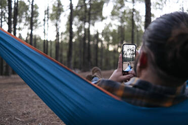 Man photographing through mobile phone lying in hammock - ASGF02177