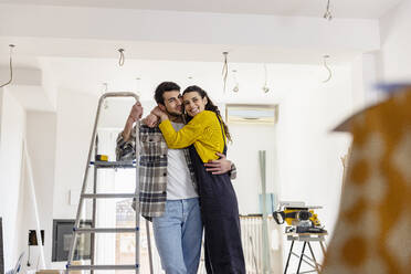 Happy woman embracing boyfriend standing by ladder in new living room - EIF03328