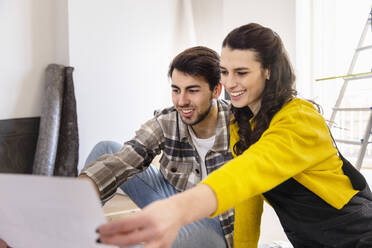 Smiling woman discussing over floor plans with boyfriend in living room - EIF03353