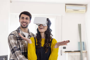 Smiling woman with VR glasses gesturing by boyfriend at new home - EIF03367