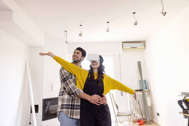 Man embracing happy girlfriend wearing VR glasses in new living room - EIF03368