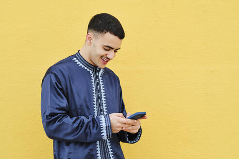 Glücklicher junger Mann, der ein Mobiltelefon benutzt und vor einer gelben Wand steht - AGOF00263