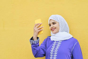 Lächelnde junge Frau, die ein Selfie vor einer gelben Wand macht - AGOF00265