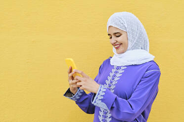 Glückliche Frau mit Mobiltelefon vor gelber Wand - AGOF00266