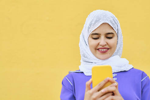 Smiling young woman surfing net through smart phone in front of yellow wall - AGOF00267