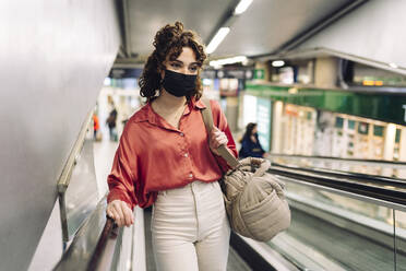 Frau mit Gesichtsschutzmaske fährt auf einer Rolltreppe im Bahnhof nach oben - JCCMF05381